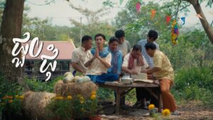 VannDa sitting on a wooden table with village elders and traditional decorations during the Back Home music video.