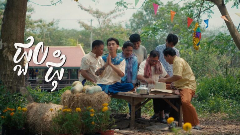VannDa sitting on a wooden table with village elders and traditional decorations during the Back Home music video.